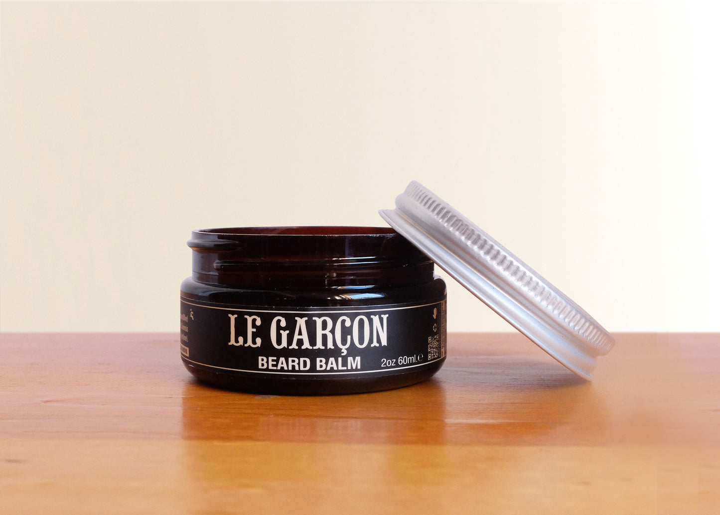 Open jar of Le Garçon Beard Balm on a wooden surface with metal lid beside it, men’s grooming product