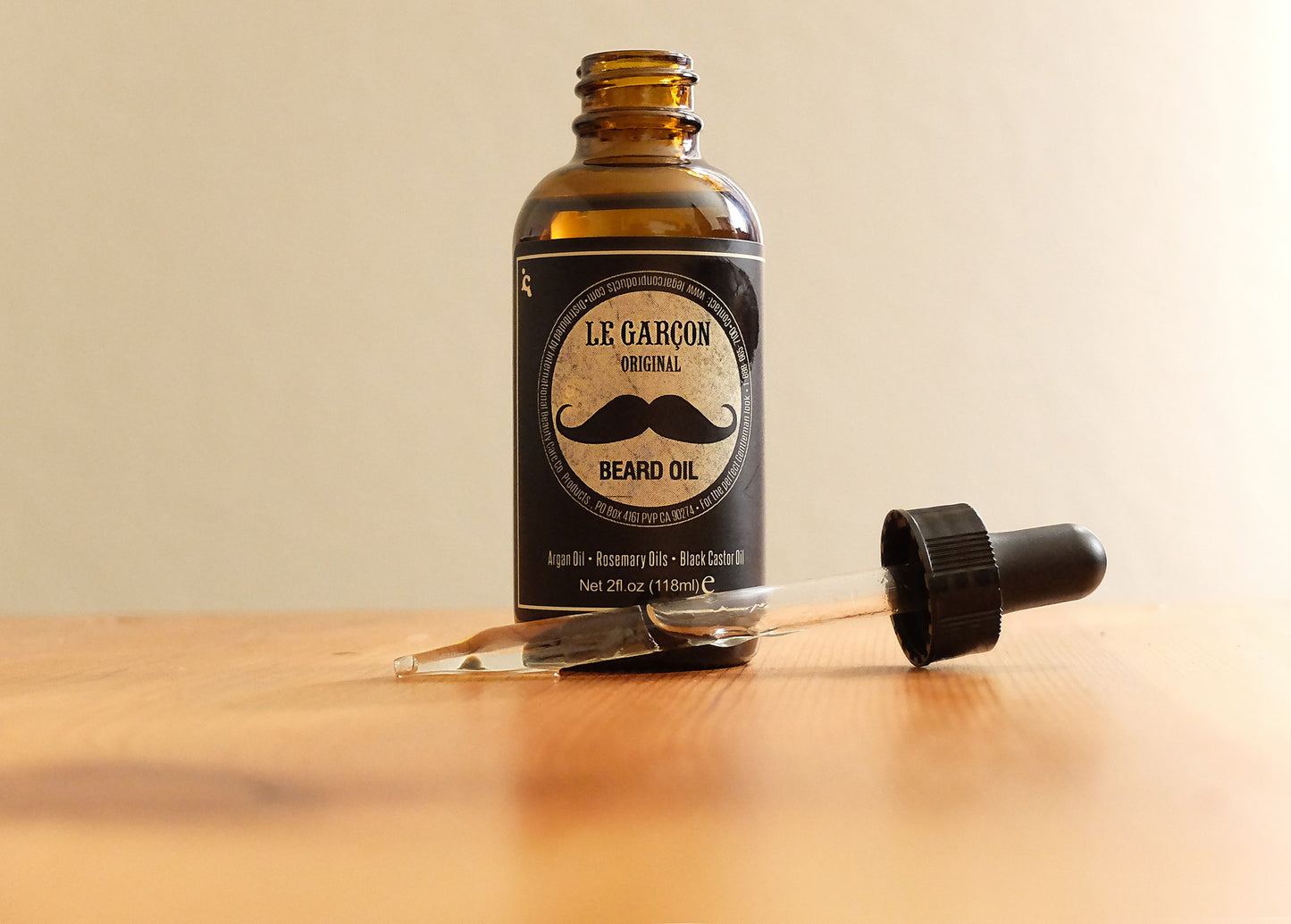 Beard oil bottle labeled 'Le Garçon Beard Oil' with dropper top, resting on a rustic wooden surface
